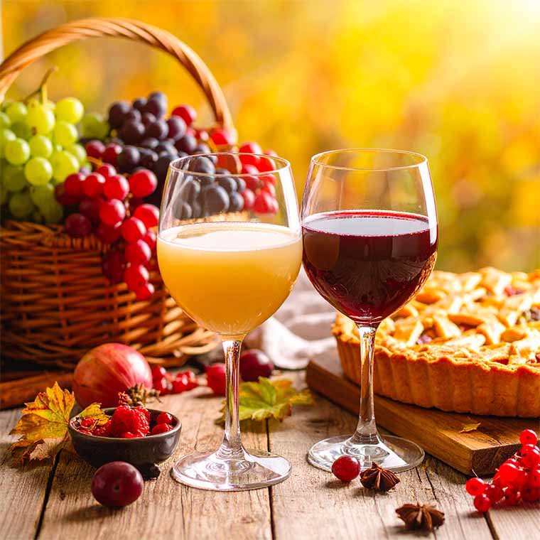 Das Foto zeigt jeweils ein Glas roten und weißen Federweißer. Daneben steht ein Flammkuchen und ein Korb mit weißen und roten Weintrauben. Alles steht auf einem rustikalen Holztisch, der herbstlich dekoriert ist.